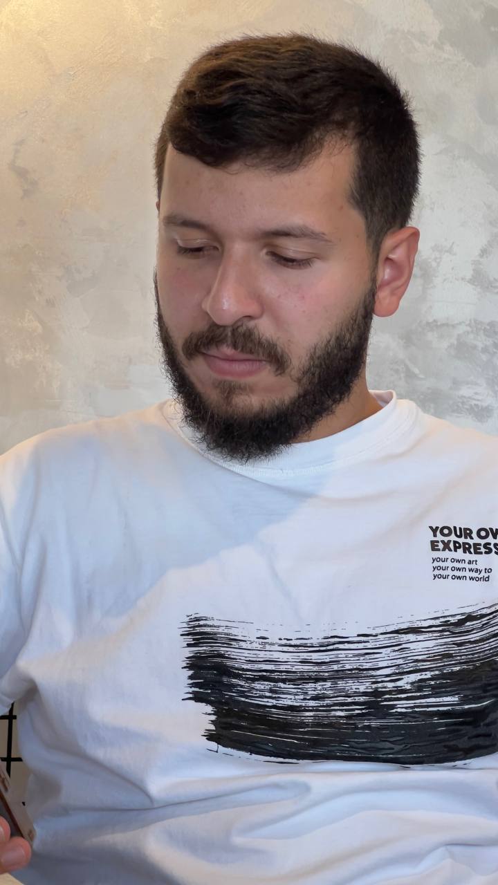 Reda, smiling softly, in a white tee against a warm plaster wall.